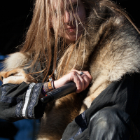 A person with long hair obscuring part of their face is holding a microphone close to their mouth. They are wearing a fur garment over a dark, intricately decorated outfit. The background is dark, with a slight glimpse of blue sky, reminiscent of the night at the 2012 Kilkim Žaibu festival. The intense expression suggests they are performing passionately.