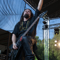 A musician with long hair and a beard, wearing a black outfit, performs on an outdoor stage at Kilkim Žaibu 2012. He holds an electric guitar and extends his right arm upwards with an open hand. Stage lights and trees are visible in the background, along with music equipment setup on the stage.