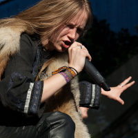 A singer with long blond hair performs passionately on stage at Kilkim Žaibu 2012, holding a microphone close to their mouth. They wear a dark top with patterned trim, leather pants, and several colorful wristbands. A fur stole drapes over their shoulders, and a dark backdrop with foliage is visible behind them.