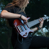 A person with long, light brown hair plays a six-string electric bass guitar at the 2012 Kilkim Žaibu festival. They are wearing a black shirt and black pants, and wristbands on both wrists. The background is an outdoor setting with trees blurred in the distance. The person seems intensely focused on the instrument.