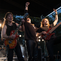 At the 2012 Kilkim Žaibu festival, three male musicians perform energetically on an outdoor stage. The one on the left plays an electric guitar, the one on the right a bass guitar, and the middle musician raises his fist. A drummer is seen in the background. Stage lights shine, and fog creates a dramatic atmosphere.