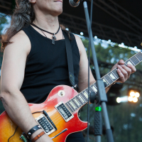 A person with long hair, wearing a black sleeveless top and wristbands, plays a sunburst electric guitar and sings into a microphone on an outdoor stage at the 2012 Kilkim Žaibu festival. The background includes stage lighting and rigging with some trees visible, creating an atmosphere that suggests a live music performance.