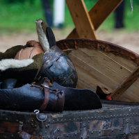A medieval helmet, fur-lined leather armor pieces, and a wooden shield rest on a weathered trunk at the 2012 Kilkim Žaibu festival. The scene is set outdoors with grass and a blurred background, suggesting a historical reenactment or period setting. Wooden beams behind the trunk form a partial structure.