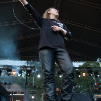 Lead singer holding a microphone and performing energetically on an outdoor stage at the 2012 Kilkim Žaibu festival. The singer is wearing a black long-sleeve shirt and black leather pants. Stage lights and amplifiers can be seen in the background, with trees visible beyond the stage structure. The scene captures a dynamic moment of the performance.