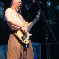 A shirtless man with a bald head and long braided ponytail passionately plays a bass guitar on stage at Kilkim Žaibu 2012. He is wearing brown, loose-fitting pants and has several accessories on his wrists. Singing or shouting into a microphone, the dark background is lit by a faint blue glow.