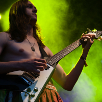 A shirtless person with long hair plays an electric guitar on stage at the 2012 Kilkim Žaibu festival, with a bright green and blue light illuminating them from behind. They are wearing a necklace and a red skirt, concentrating intensely on their performance.