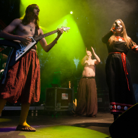 On a dimly lit stage at the 2012 Kilkim Žaibu festival, a barefoot guitarist in a long skirt plays next to a singer in a black and red traditional-style dress holding a microphone. In the background, a shirtless man in a long skirt passionately claps his hands. A drum set is visible on the left side of the stage.