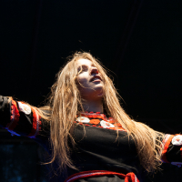 A person with long, flowing hair stands on a dimly lit stage at the 2012 Kilkim Žaibu festival, with arms outstretched. They are wearing a black garment adorned with red and white floral patterns and a red belt. The lighting casts dramatic shadows, emphasizing their expressive pose against the dark background.