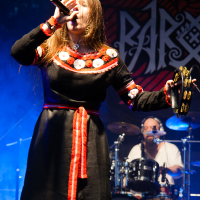 A woman in a black dress with red and white embroidery sings passionately into a microphone on stage at Kilkim Žaibu 2012. Her long hair flows over her shoulders as she holds a tambourine in her left hand. A drummer performing in the background is partially visible, with stage lights creating a blue illumination around them.