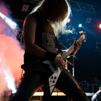 A guitarist with long hair plays an electric guitar energetically on a dimly lit stage at Kilkim Žaibu 2012. He is wearing a sleeveless black shirt and black leather pants. Smoke and colorful stage lights create a dramatic atmosphere. The guitar has a white body, and his hair obscures his face as he leans forward.