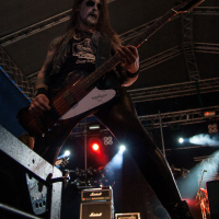 A rock musician with a painted face and long hair plays an electric bass guitar onstage at the 2012 Kilkim Žaibu festival. He wears a sleeveless shirt and tight black pants, standing with legs apart under a large stage with dramatic lighting and visible rigs and amplifiers behind him.