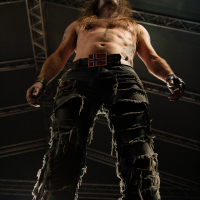 A muscular, shirtless man with long hair stands under stage lighting, seen from a low angle. He wears tattered black pants, black wristbands, and a belt adorned with a flag resembling Norway's. Amidst the backdrop of metal framework and stage equipment at Kilkim Žaibu 2012, he appears deep in performance.