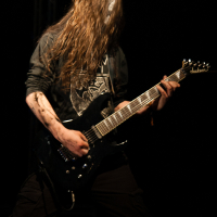 A musician with long, flowing hair obscuring their face is playing an electric guitar with intensity at the Kilkim Žaibu festival. Dressed in dark clothing and deeply focused on their performance on stage against a dark background, they wield a black and white designed guitar, capturing the essence of 2012.