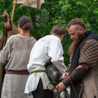 Three people, dressed in medieval-style clothing, stand outdoors in a forested area at Kilkim Žaibu 2012. One person on the left holds a flag with a design and wears a tunic. The person in the center is adjusting a piece of armor, and the person on the right has a long beard and is wearing quilted armor.
