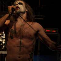 At the Kilkim Žaibu festival in 2012, a shirtless man with long hair performs on stage, holding a microphone to his mouth with one hand. His face is painted white with dark makeup around his eyes and mouth. He has tattoos, including an inverted cross on his torso. The background is dark and filled with stage equipment.