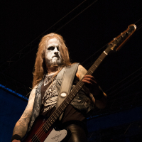 A musician with long hair and face paint plays a bass guitar on a dimly lit stage at Kilkim Žaibu festival in 2012. He wears a sleeveless denim vest over a black shirt with intricate designs. The background is dark, with subtle stage lighting hinting at a live performance setup.