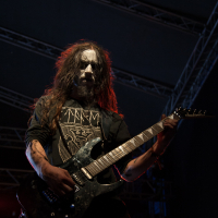 A person with long hair and face paint, reminiscent of a black metal style, is playing an electric guitar on stage at Kilkim Žaibu 2012. They are wearing a black shirt with a geometric print. The background is dark, and the stage is lit with overhead lights, emphasizing the intense atmosphere of the performance.