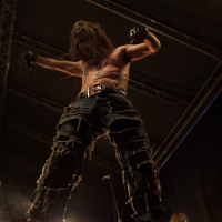 At the 2012 Kilkim Žaibu festival, a shirtless performer on stage stands with arms raised, holding a microphone. He wears distressed, ripped jeans with a belt and fingerless gloves. His long hair obscures his face. Stage lights illuminate him from below, highlighting the dramatic pose and creating an intense atmosphere.
