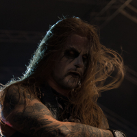 A man with long, flowing hair and heavy black and white face paint stands under stage lighting at the 2012 Kilkim Žaibu festival, arms crossed. He wears a sleeveless outfit, showcasing his tattoo-covered arms, and has a serious expression with a distant gaze. The background is dark, with indistinct lighting structures.