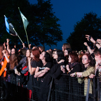 A crowd of excited people at an outdoor concert at dusk, many with raised arms, some holding flags in the background. They are standing behind a metal barrier with trees silhouetted against a darkening sky behind them. The atmosphere is lively and energetic, reminiscent of Kilkim Žaibu 2012.