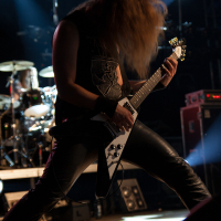 A musician with long hair in motion plays an electric guitar on stage at the 2012 Kilkim Žaibu festival, wearing a sleeveless shirt and black leather pants. In the background, a drummer plays a drum set. The stage is illuminated by vibrant blue and white lights, creating a dynamic atmosphere.