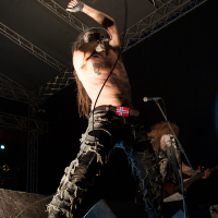 A shirtless musician with long hair performs on stage at Kilkim Žaibu 2012, wearing torn black jeans and a belt with a Norwegian flag buckle. He has white face paint with black details and energetically raises his arms, holding a microphone. Another band member plays guitar in the background under stage lights.