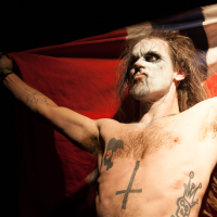 A shirtless person with long hair and face paint stands with arms raised, holding a red and black flag behind them. They have tattoos, including a large cross on their torso and various others on their arms and chest. The dark background highlights their dramatic appearance, resembling the fervor of the Kilkim Žaibu festival.
