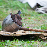 A medieval-style helmet and a wooden-handled weapon, possibly a mace or axe, rest on a makeshift wooden surface outdoors with greenery in the background. The scene suggests a historical reenactment or display of medieval artifacts at Kilkim Žaibu 2012, with a soft focus on the background elements.