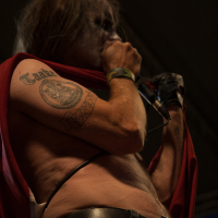 A singer with long hair and face paint holds a microphone close to his mouth. He is shirtless, revealing a tattoo on his right bicep. Wearing a red cape and a belt with a buckle displaying the Norwegian flag, he performs at the 2012 Kilkim Žaibu festival. The dark background suggests an indoor or nighttime performance.