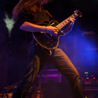 At Kilkim Žaibu 2012, a guitarist with long hair performs energetically on stage, illuminated by dramatic lighting. He plays a black electric guitar and is dressed in a black shirt and blue jeans. Amplifiers, including one from Marshall, are visible in the background, surrounded by blue and purple stage smoke.