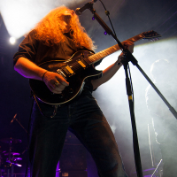 A musician with long, wavy blond hair and a beard performs on stage with a black electric guitar. Wearing a black shirt and blue jeans, he stands under dramatic stage lighting, casting a smoky, atmospheric glow. A microphone is positioned in front of him amid band equipment—evoking the raw energy of Kilkim Žaibu 2012.
