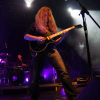 A guitarist with long hair covering their face is passionately playing an electric guitar on a dimly lit stage at the 2012 Kilkim Žaibu festival. The musician, dressed in dark clothes, stands near a microphone while a drummer can be seen in the background, both illuminated by purple and white stage lights.
