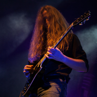 A person with long, flowing hair plays an electric guitar on stage in a dimly lit, smoky environment at Kilkim Žaibu festival. They wear a dark shirt with the sleeves rolled up and blue jeans. The scene is filled with dramatic lighting, highlighting the intense focus and dynamic presence of the musician.