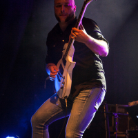 A bald person is energetically playing an electric guitar on stage at the 2012 Kilkim Žaibu festival, illuminated by dramatic concert lighting. They are wearing a dark shirt and jeans, fully engaged in the performance. A spotlight shines from the lower left, casting dynamic shadows against a dim and blurred background featuring indistinct equipment.