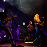 At the Kilkim Žaibu 2012 festival, a rock band performs energetically on a dimly lit stage with purple and blue lighting. The lead guitarist with long hair plays passionately while three other band members play guitar, bass, and keyboard. A faint image of a winged figure is visible on the backdrop.