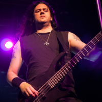 A musician with long black hair and tattoos plays an electric bass guitar on stage at the 2012 Kilkim Žaibu festival. He is illuminated by purple stage lights and wears a sleeveless black shirt, a necklace with a cross, and a wristband. The background includes stage equipment and lighting fixtures.