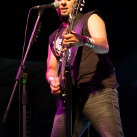 A bald musician with a beard is passionately playing an electric guitar on stage at Kilkim Žaibu 2012. He is wearing a sleeveless black shirt, gray jeans, and metal bracelets. A microphone is positioned in front of him, and the stage is dimly lit with purple and yellow spotlights.