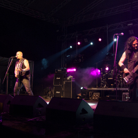 At the 2012 Kilkim Žaibu festival, a rock band performs live on stage at night, illuminated by vibrant purple and white lights. The lead singer, in a sleeveless shirt, plays guitar and sings into the mic. To his right, a long-haired bassist is engrossed in playing. Large speaker stacks and amplifiers surround them.