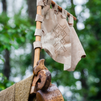A tattered, ancient-looking beige flag with a geometric design hangs from a wooden pole adorned with leather straps. Below the flag, there is a wooden drinking vessel with intricate carvings. Set against the backdrop of a dense green forest, this scene evokes memories of Kilkim Žaibu 2012 festival.