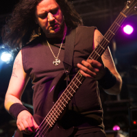 A musician with long dark hair is playing an electric bass guitar on stage at the 2012 Kilkim Žaibu festival. He is wearing a sleeveless black shirt, black wristbands, and a necklace with a pendant. The stage is lit with colorful lights, including purple and blue, creating a vibrant atmosphere.