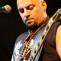 A bald, bearded musician wearing a sleeveless shirt and beaded necklaces is playing an electric guitar on stage at Kilkim Žaibu 2012. He is looking down at his guitar while performing. A microphone is visible to his left. The background is dark and out of focus, highlighting the performer.