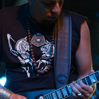 A bald man intensely plays an electric guitar at the 2012 festival. He wears a black sleeveless shirt with a graphic of two stylized figures, a large pendant, and multiple beaded necklaces. He has several rings and bracelets on both hands, set against a blurred background with blue lighting effects.