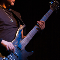 A man wearing a sleeveless black shirt and a chain necklace plays an electric bass guitar on stage at the 2012 Kilkim Žaibu festival. He focuses intently on the instrument, with his left hand positioned on the fretboard and his right hand plucking the strings. The dark background highlights him under stage lighting.