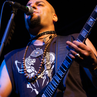 A bald musician with a goatee is playing an electric guitar and singing into a microphone at the 2012 Kilkim Žaibu festival. He is wearing a sleeveless black shirt with a graphic design and layered necklaces, including beads and an amulet. The dark background highlights him with stage lighting.