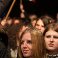 At the 2012 Kilkim Žaibu festival, a blonde woman in a green jacket stands among a dense crowd, looking ahead with a thoughtful expression. Another young woman behind her has long dark hair and a neutral look. The crowd appears focused, and in the background, someone holds up a red and white flag.