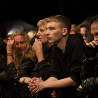 A group of concertgoers, predominantly young adults, is closely packed together behind a barricade at an outdoor nighttime event. They are attentively watching the stage, dressed in dark casual attire, some with long hair and others with short hair. The atmosphere at the 2012 Kilkim Žaibu festival suggests anticipation and excitement.