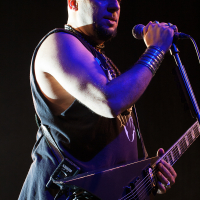 A bald musician with a goatee passionately sings into a microphone while playing a V-shaped electric guitar at Kilkim Žaibu 2012. He wears a sleeveless black shirt, and his wrists are adorned with multiple metal bracelets. The festival stage lighting highlights his arms and face against a dark background.