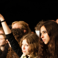 A crowd of people at a festival, mostly young adults, some with long hair. A woman in the foreground has dark, straight hair and a serious expression, while another woman nearby, with curly hair, raises her arm excitedly. The background is dark, highlighting the individuals' faces with Kilkim Žaibu 2012 concert lighting.