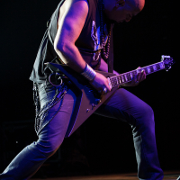 A bald musician wearing a sleeveless black shirt, jeans, and boots is passionately playing an angular electric guitar on stage at Kilkim Žaibu 2012. He is leaning forward with one leg bent, creating an intense and dynamic pose. Chains and wristbands add to his rock or heavy metal aesthetic. The background is dark.