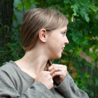 A woman with light brown hair braided over one shoulder stands outdoors at the 2012 Kilkim Žaibu festival, looking to her left. She wears a loose-fitting, long-sleeved, grayish-brown tunic. The background is lush with green foliage, indicating a natural, wooded area. She is holding her braid with both hands.